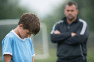 Criança abaixa a cabeça após errar em jogo de futebol infantil enquanto o treinador observa ao fundo